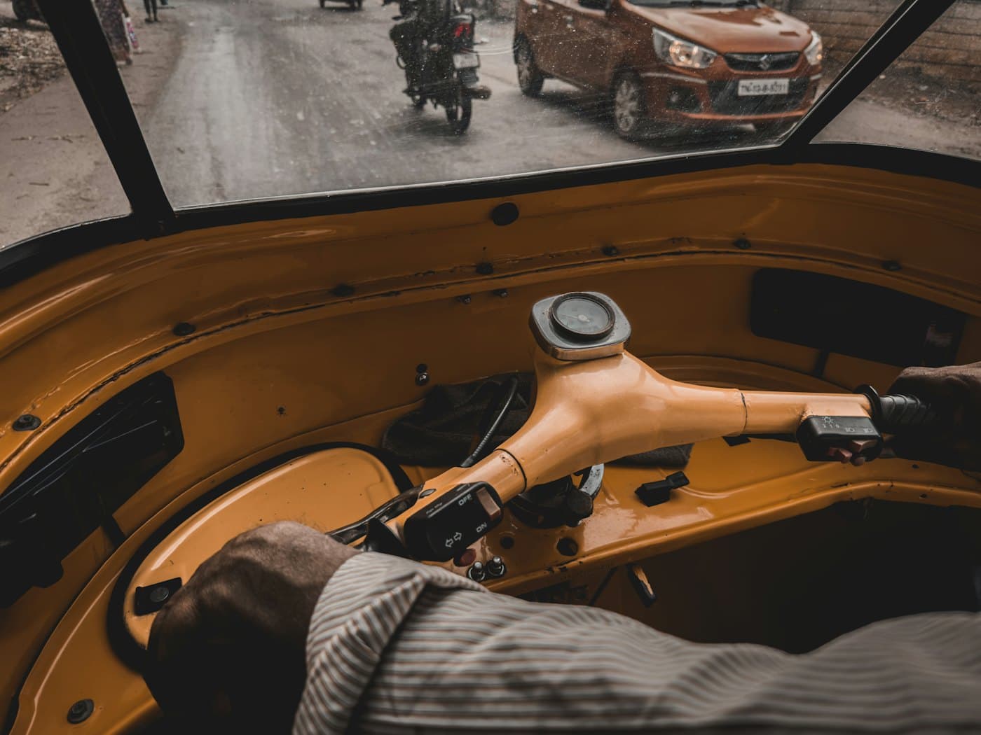 A driver's hand on an auto-rickshaw handlebar, the street ahead through the windscreen