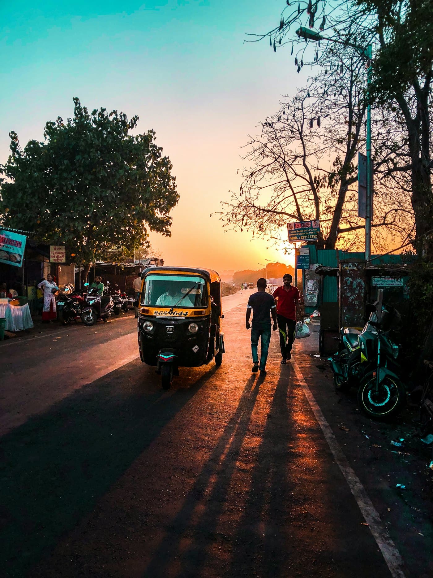 An auto-rickshaw on a tree-lined street at sunset, people walking alongside